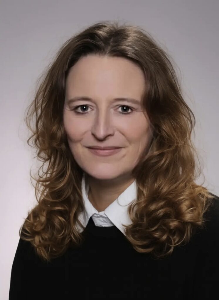 Porträt einer Frau mit schulterlangem, lockigem Haar vor neutralem Hintergrund.