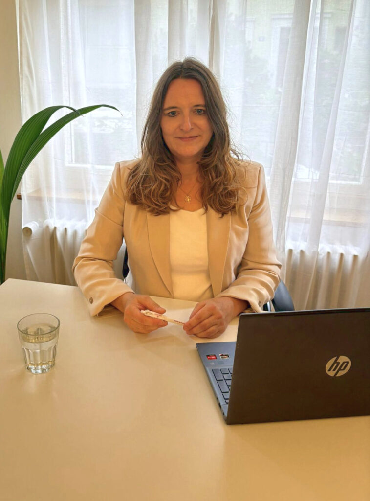 Frau sitzt an einem Schreibtisch mit Laptop, Glas Wasser und Stift in ruhiger Arbeitsumgebung.